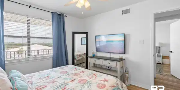 Image 3: Bright bedroom featuring a large window with horizontal blinds, a ceiling fan with integrated lighting, wood-finish flooring, and white walls