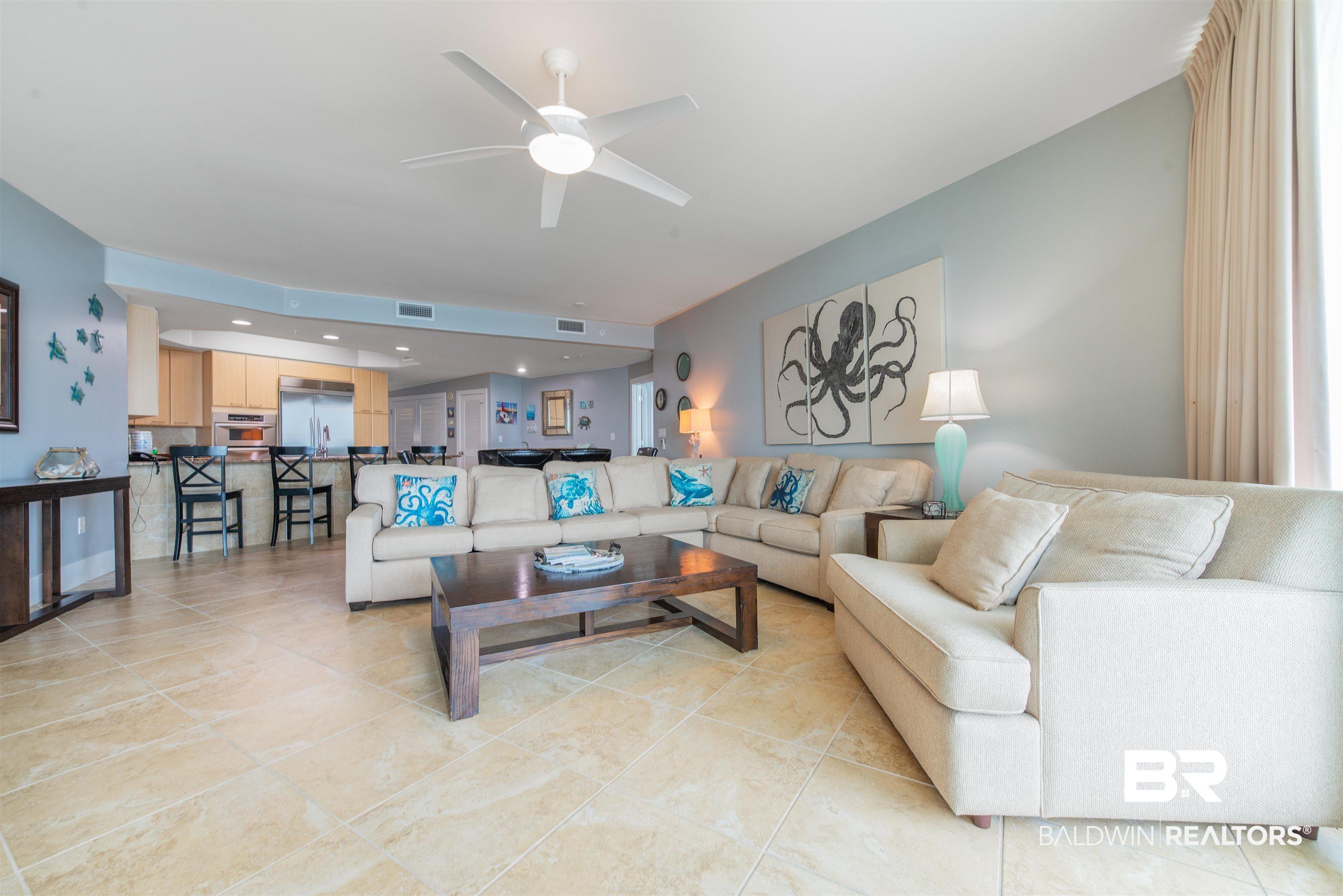 Image 3: Open-concept living space featuring light-toned tile flooring, a recessed ceiling with a white ceiling fan, and a kitchen area with light wood cabinetry and stainless steel appliances