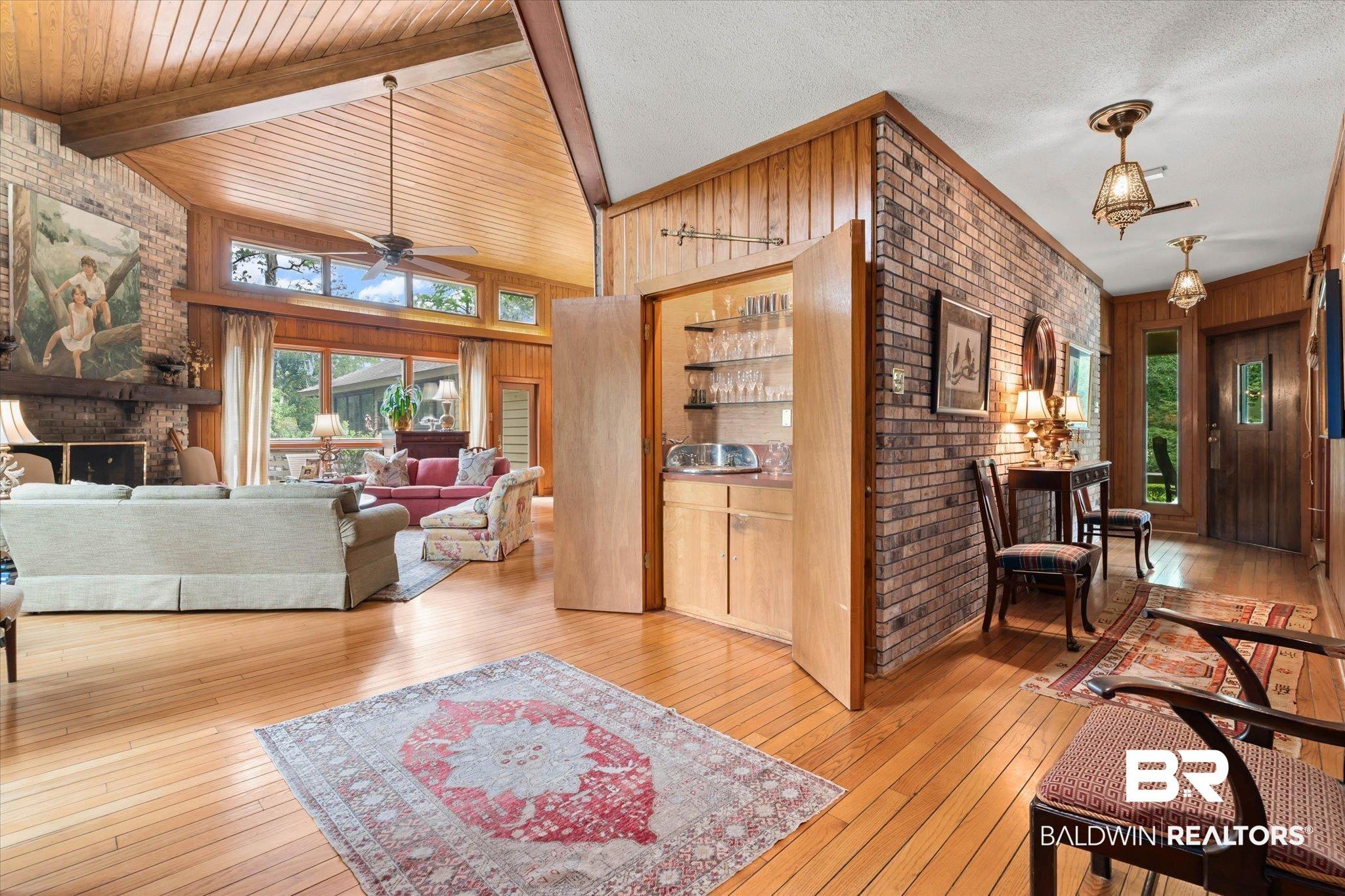 Image 4: Spacious living area featuring a vaulted wood-paneled ceiling with exposed beams, wood-finish flooring, a brick fireplace, and a built-in wet bar with glass shelving