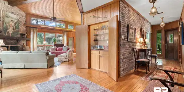 Image 4: Spacious living area featuring a vaulted wood-paneled ceiling with exposed beams, wood-finish flooring, a brick fireplace, and a built-in wet bar with glass shelving