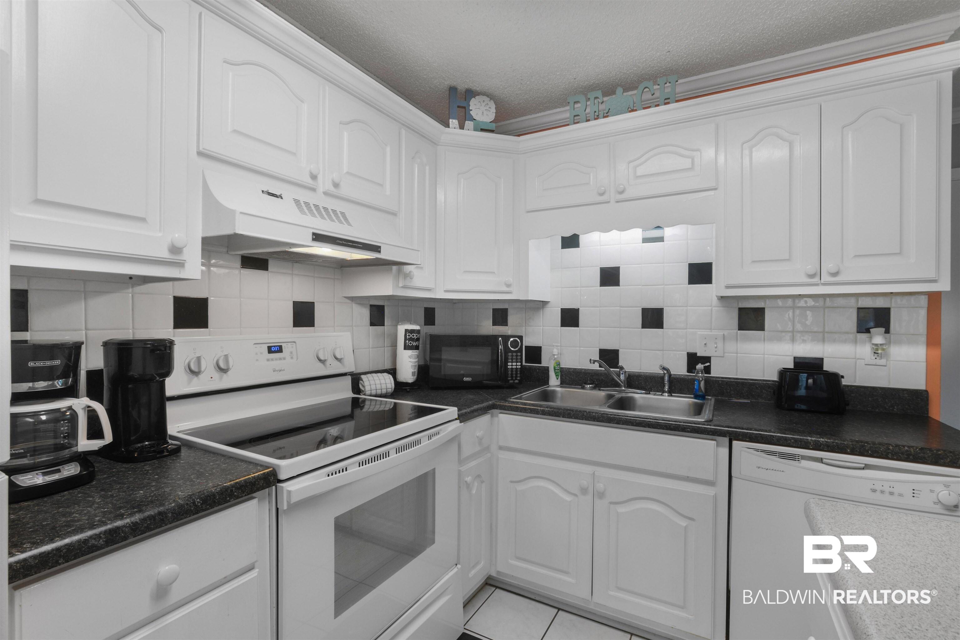 Image 4: Kitchen featuring white cabinetry, black speckled countertops, a white tile backsplash with black accent squares, a double basin stainless steel sink, and a built-in range with an overhead vent