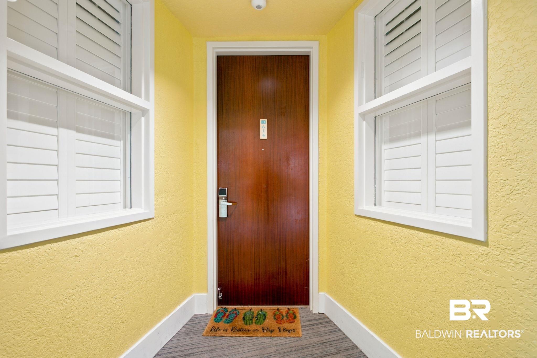 Image 2: Entryway featuring a rich wood-finish door with modern digital lock, flanked by two white-framed windows with louvered shutters, all set against textured yellow walls with white baseboards and wood-finish flooring