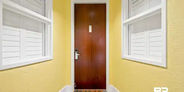 Image 2: Entryway featuring a rich wood-finish door with modern digital lock, flanked by two white-framed windows with louvered shutters, all set against textured yellow walls with white baseboards and wood-finish flooring