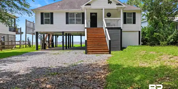 Image 4: Elevated coastal-style residence featuring white siding, a dark shingle roof, and black window shutters