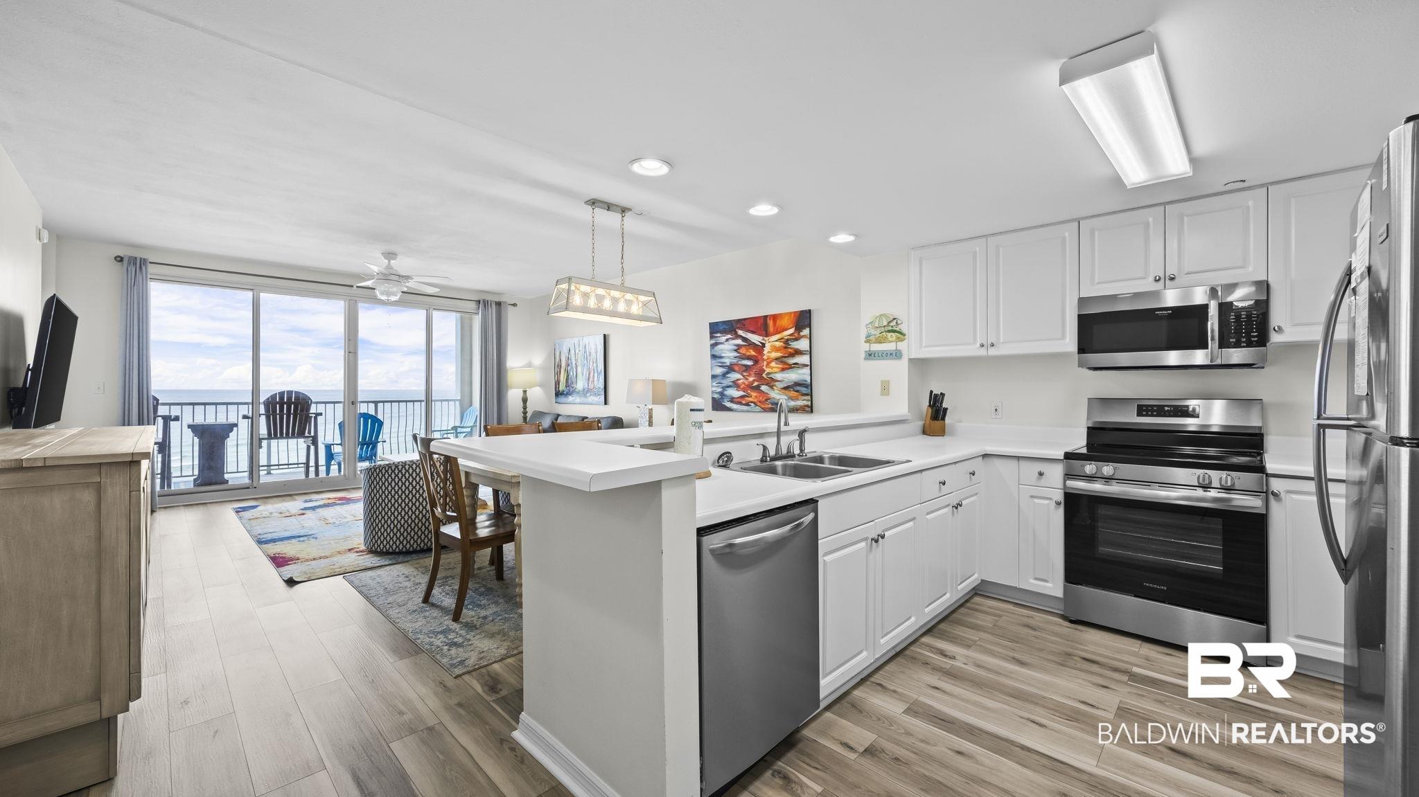 Image 4: Open-concept living area with wood-finish flooring, white cabinetry, stainless steel appliances, and a balcony with waterfront scenery