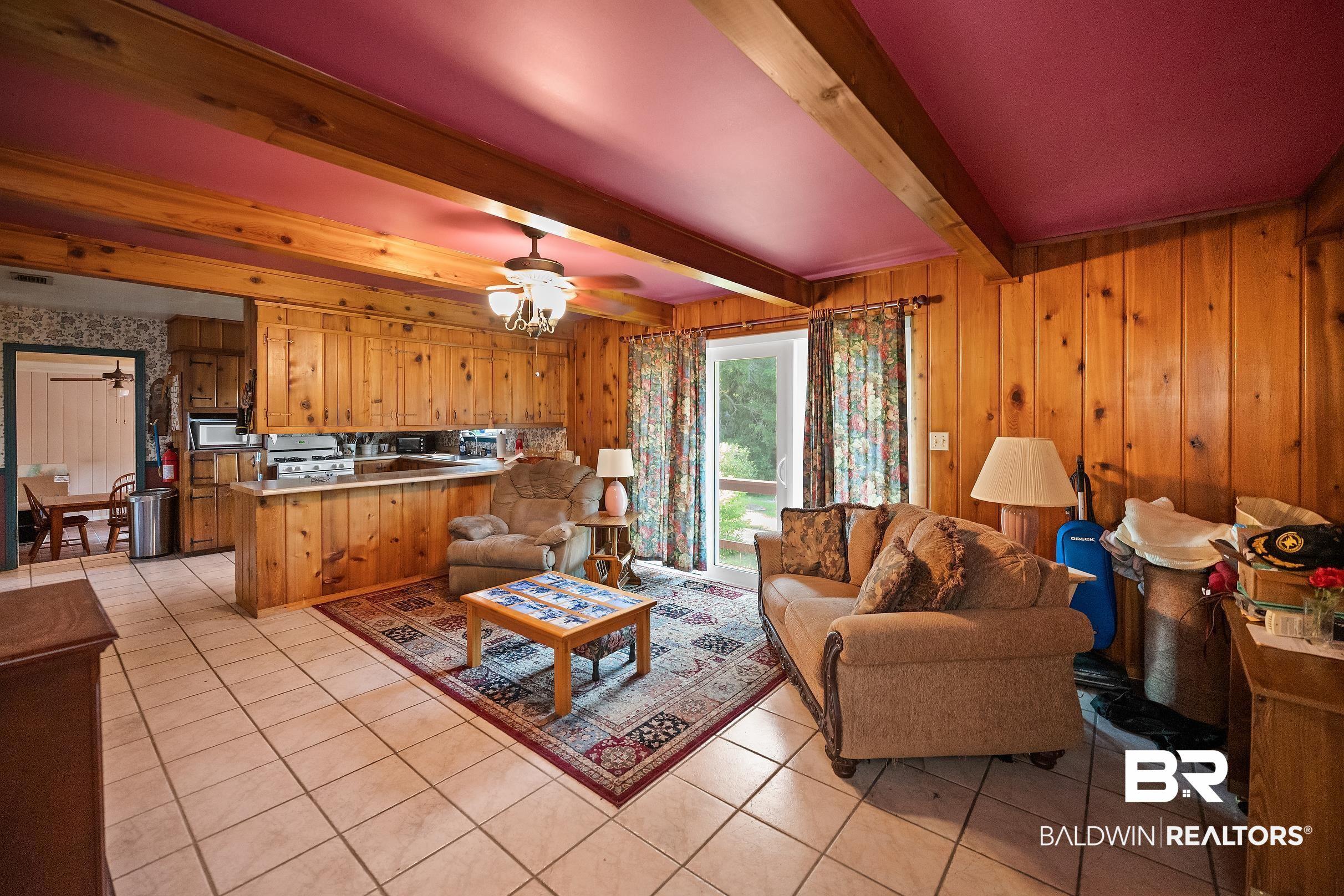 Image 4: Open concept living area featuring wood paneling, tiled flooring, exposed wood beams, and a ceiling fan with integrated lighting
