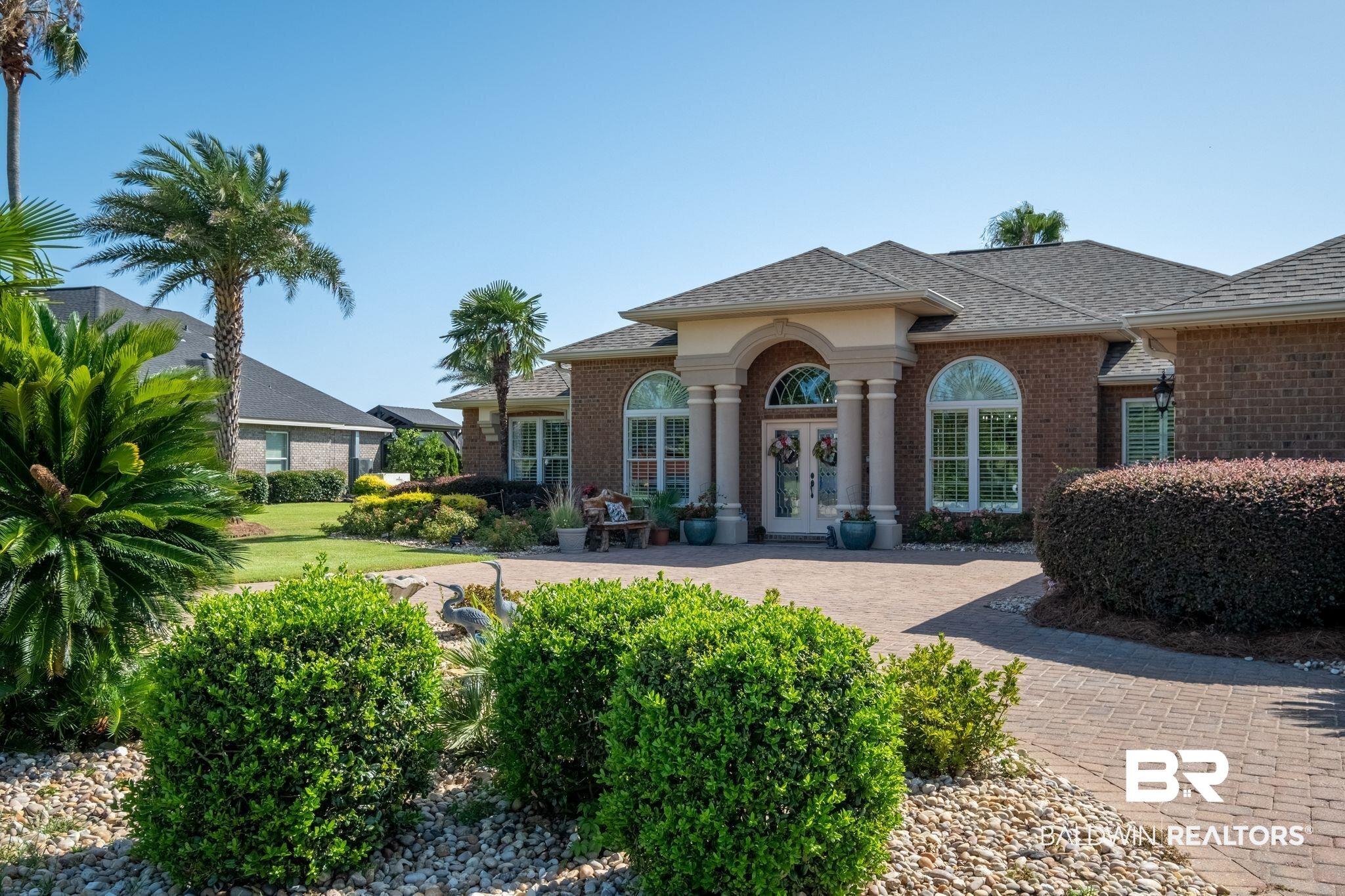 Image 3: Brick facade residence featuring a covered entrance with columns, arched windows, a paver driveway, and mature landscaping with palm trees