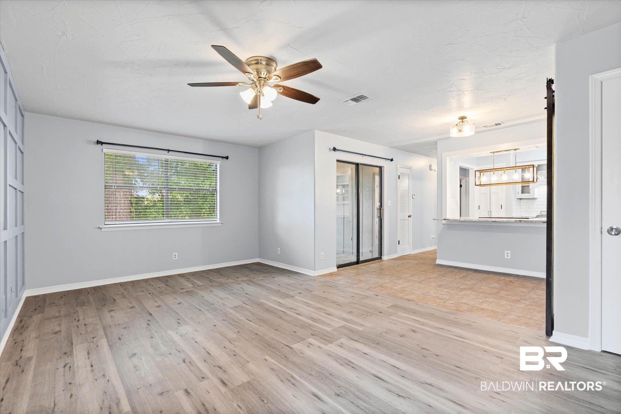 Image 3: Spacious living area featuring wood-finish flooring, a ceiling fan with integrated lighting, and a large window with blinds