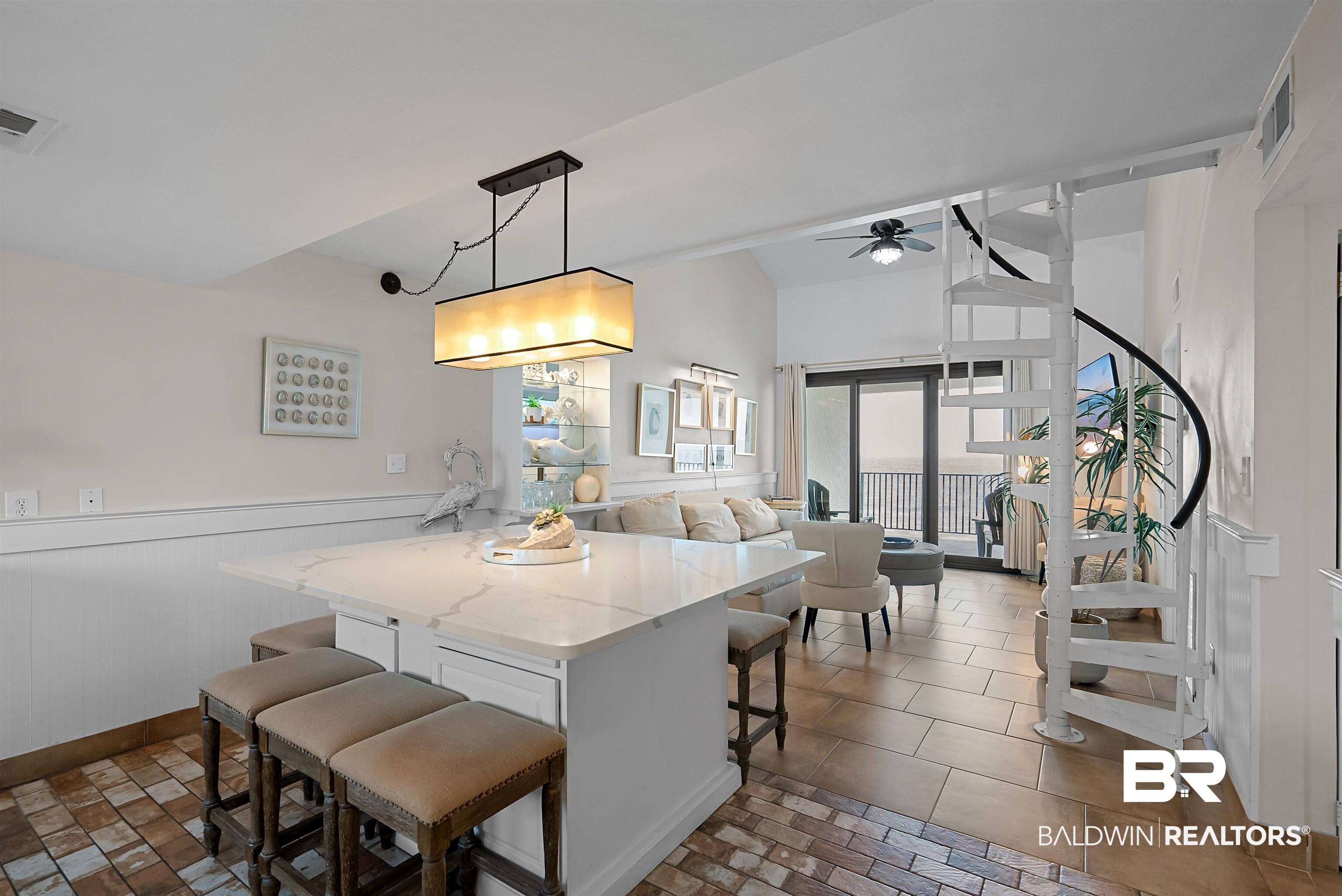 Image 2: Open-concept living space with a kitchen island featuring a light-toned countertop, a linear pendant light fixture, and tile flooring