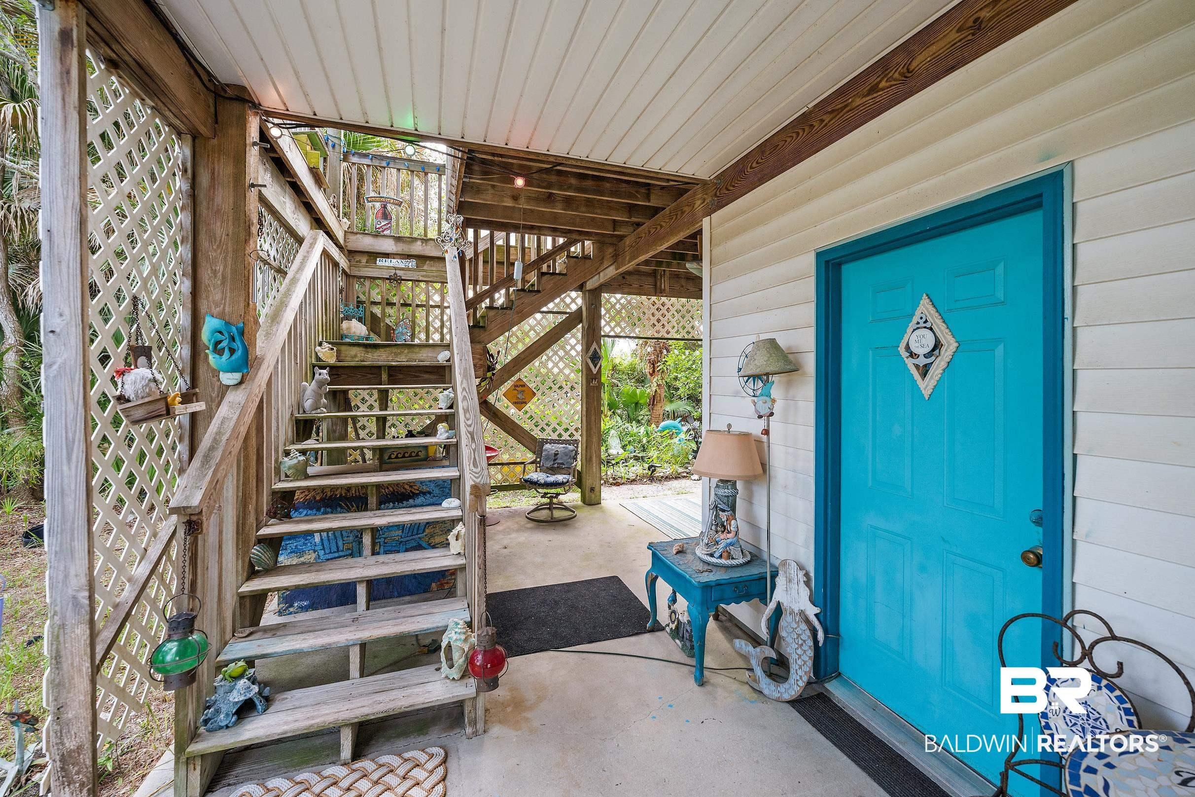 Image 3: Covered concrete patio area featuring a vibrant teal entry door, white horizontal siding, and a prominent wood staircase with lattice accents