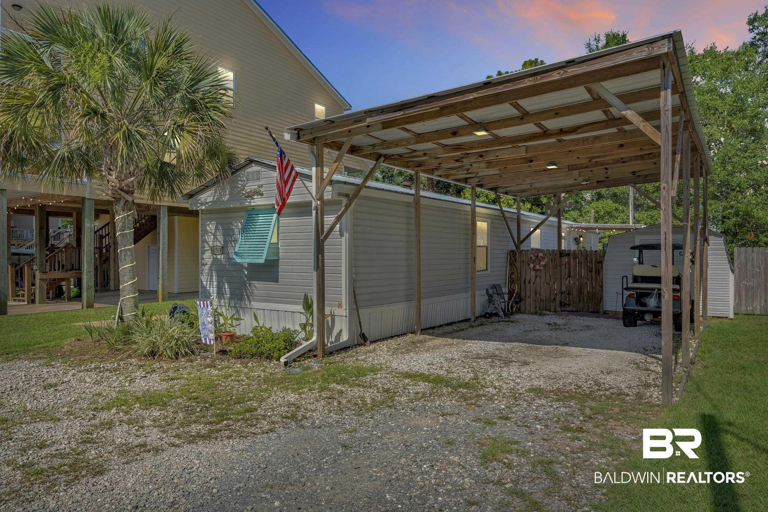 Image 3: Gravel parking area featuring a covered carport structure, an adjacent storage shed with horizontal siding, and a partial view of a multi-story residence with exterior stairs