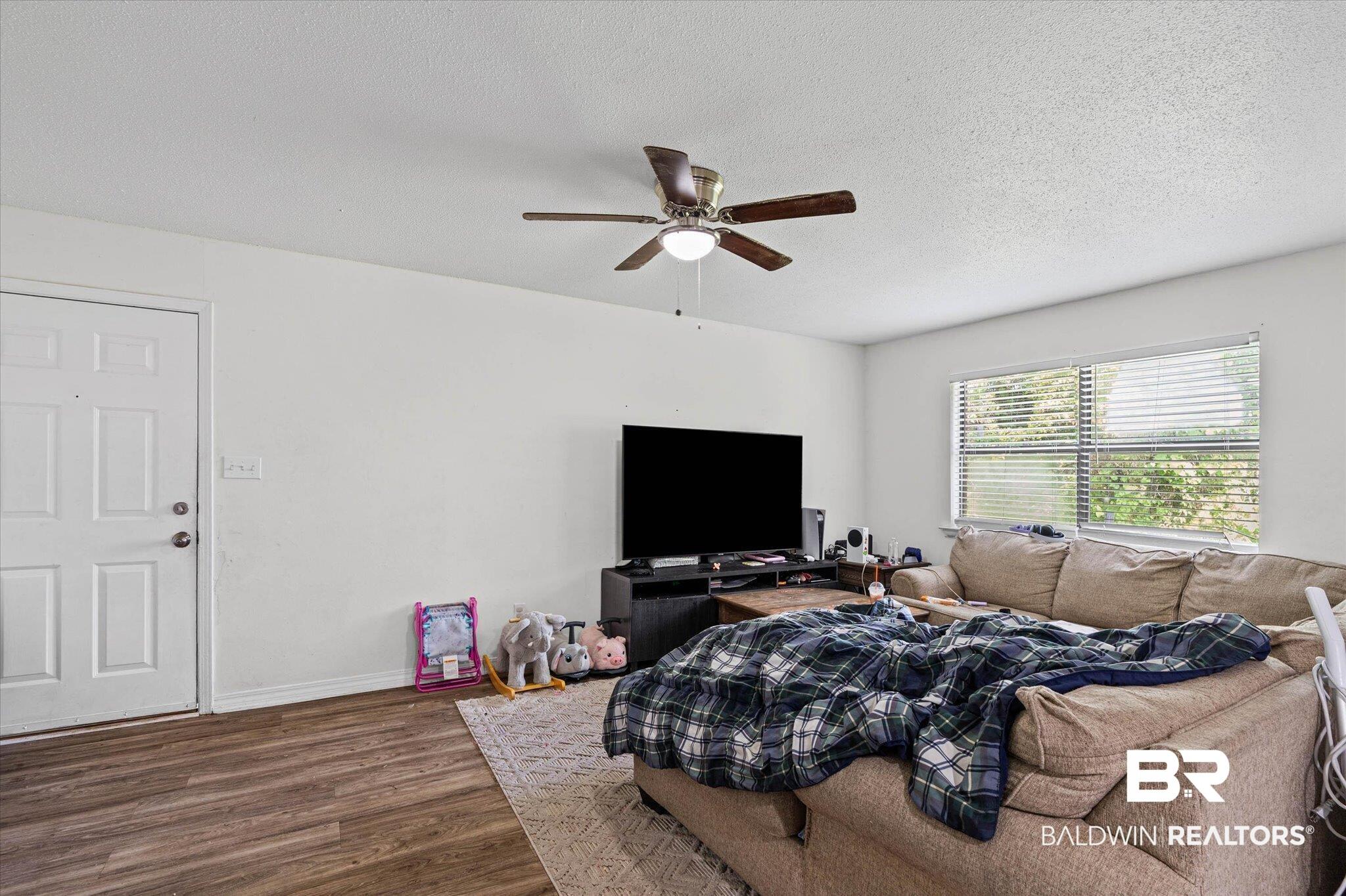 Image 2: Living area featuring wood-finish flooring, a white paneled door, a ceiling fan with integrated lighting, and a window with blinds