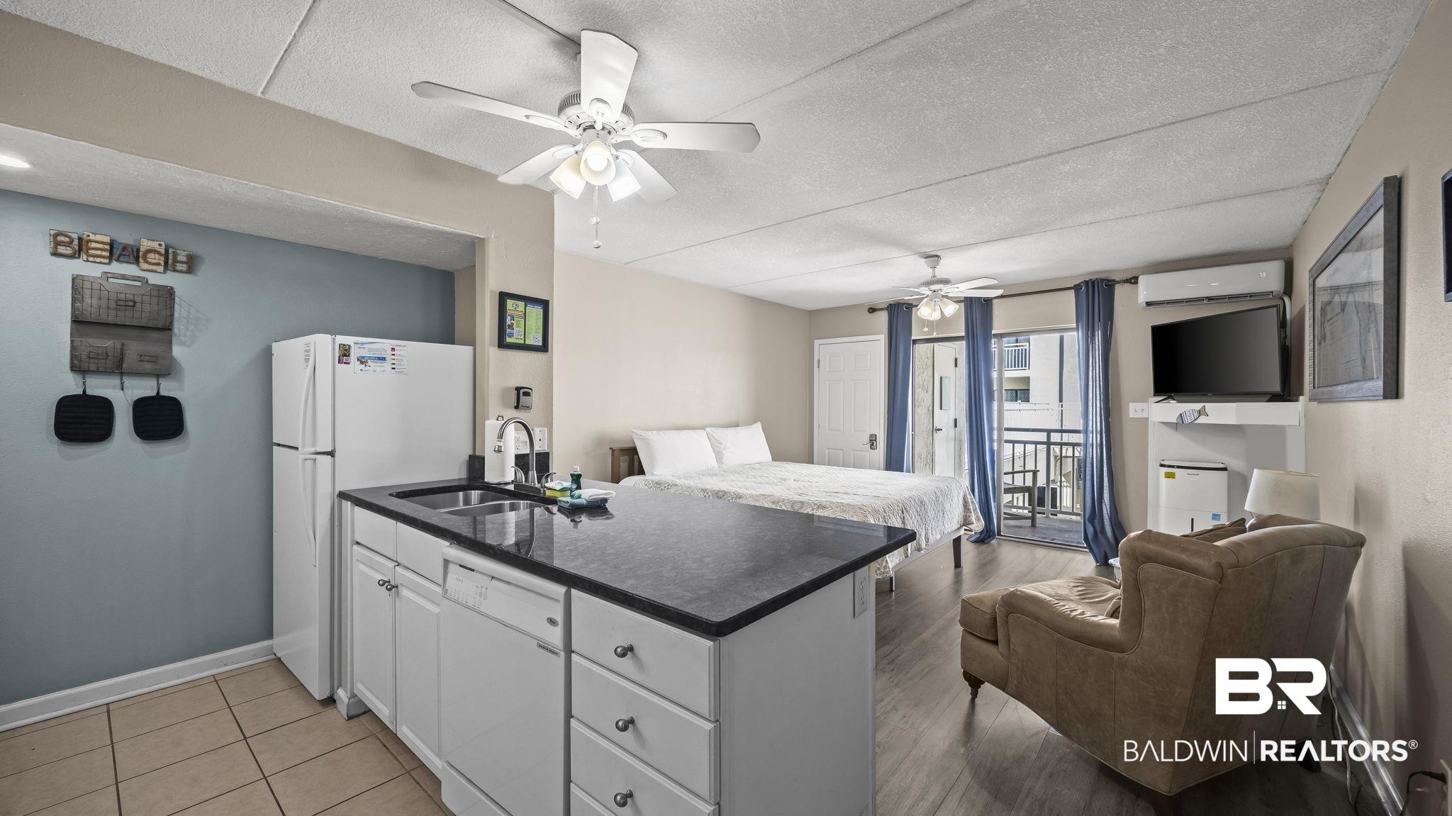 Image 4: Open-concept living area featuring wood-finish flooring, a kitchen island with a dark countertop and white cabinetry, a built-in refrigerator, and a ceiling fan with integrated lighting