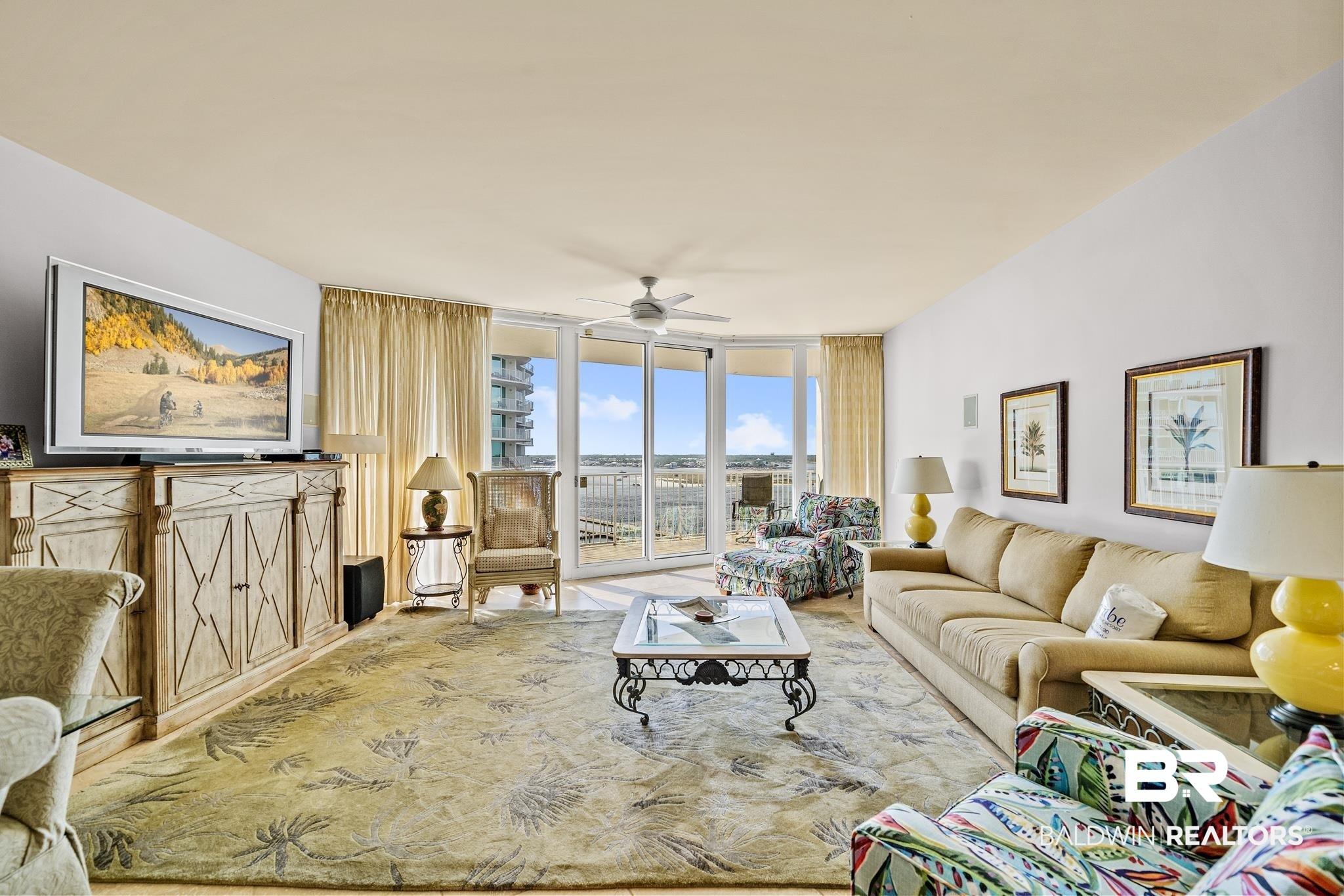 Image 2: Spacious living area featuring large sliding glass doors, balcony access, a ceiling fan, light-toned walls, and decorative wall art