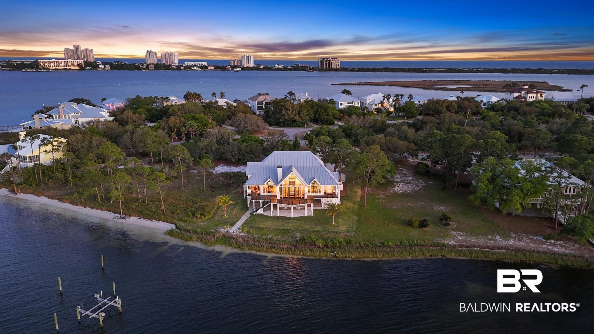 Image 2: Waterfront property featuring a prominent residence with a large elevated deck, a white sand shoreline, a private dock structure, and mature trees encompassing the lot