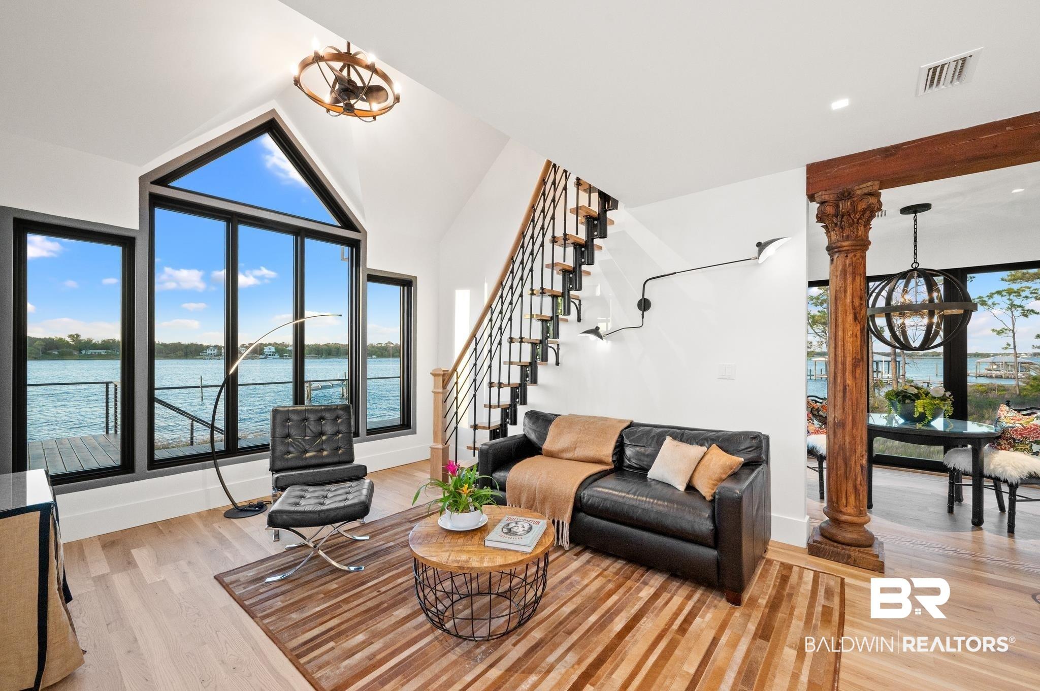Image 4: Spacious living area featuring expansive windows with water scenery, soaring vaulted ceilings, wood-finish flooring, a contemporary open-tread staircase with metal balusters, and a decorative wooden column