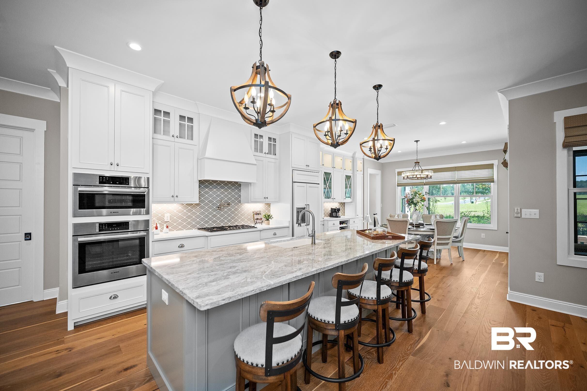 Image 4: Spacious kitchen featuring an oversized island with countertop seating, wood-finish flooring, white cabinetry, a built-in double oven, and stainless steel appliances