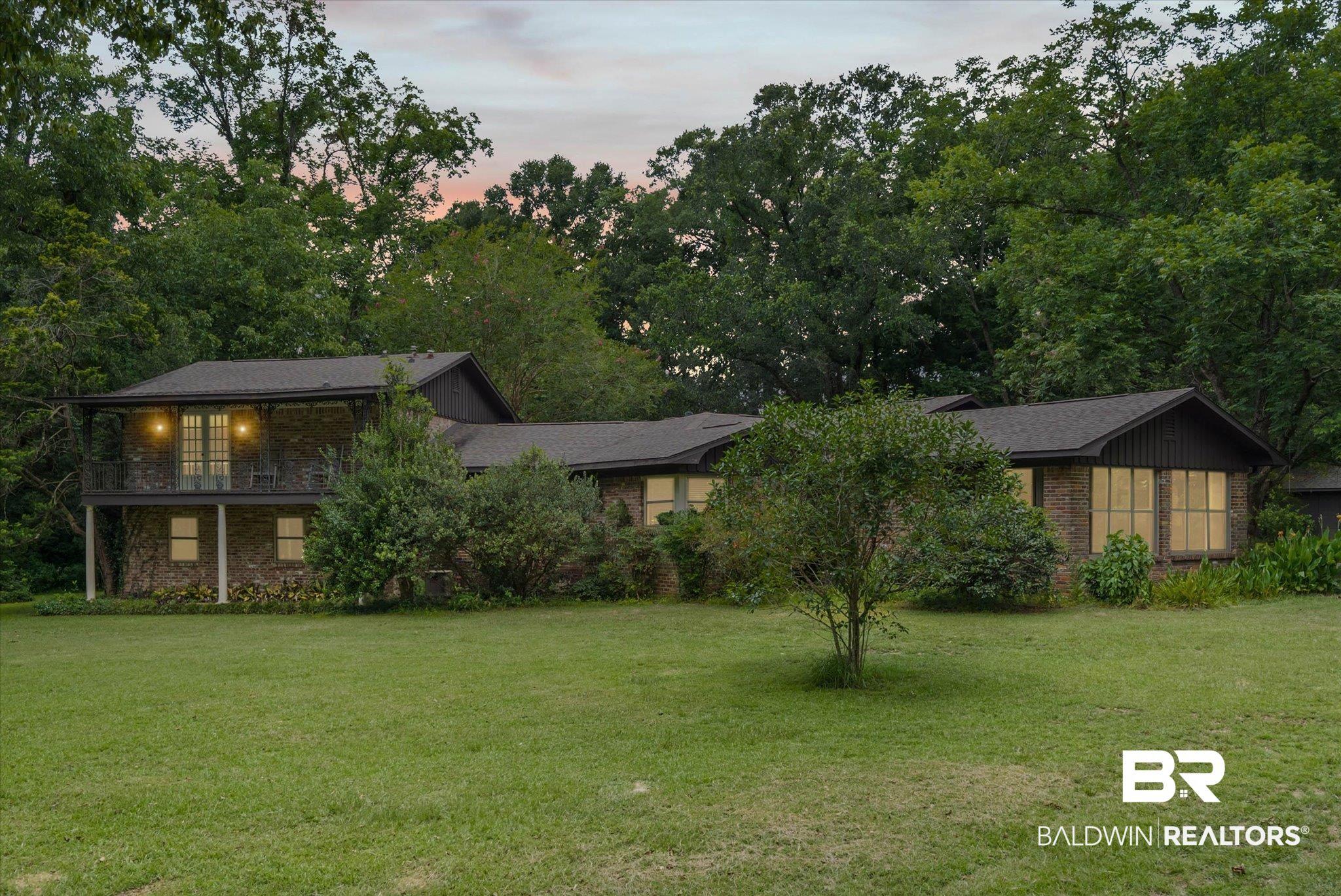 Image 1: Brick facade residence featuring a second-story balcony with metal railing, multiple windows, and a surrounding green landscape with mature trees