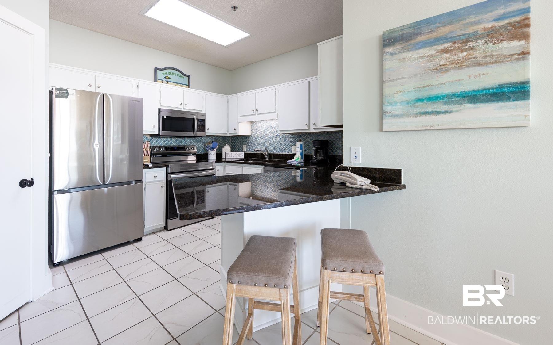 Image 4: Kitchen featuring white cabinetry, stainless steel appliances, dark stone countertops, a blue mosaic tile backsplash, and white tiled flooring