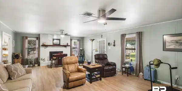 Image 4: Spacious living area featuring wood-finish flooring, a white-painted brick fireplace with a wood mantle, and two ceiling fans