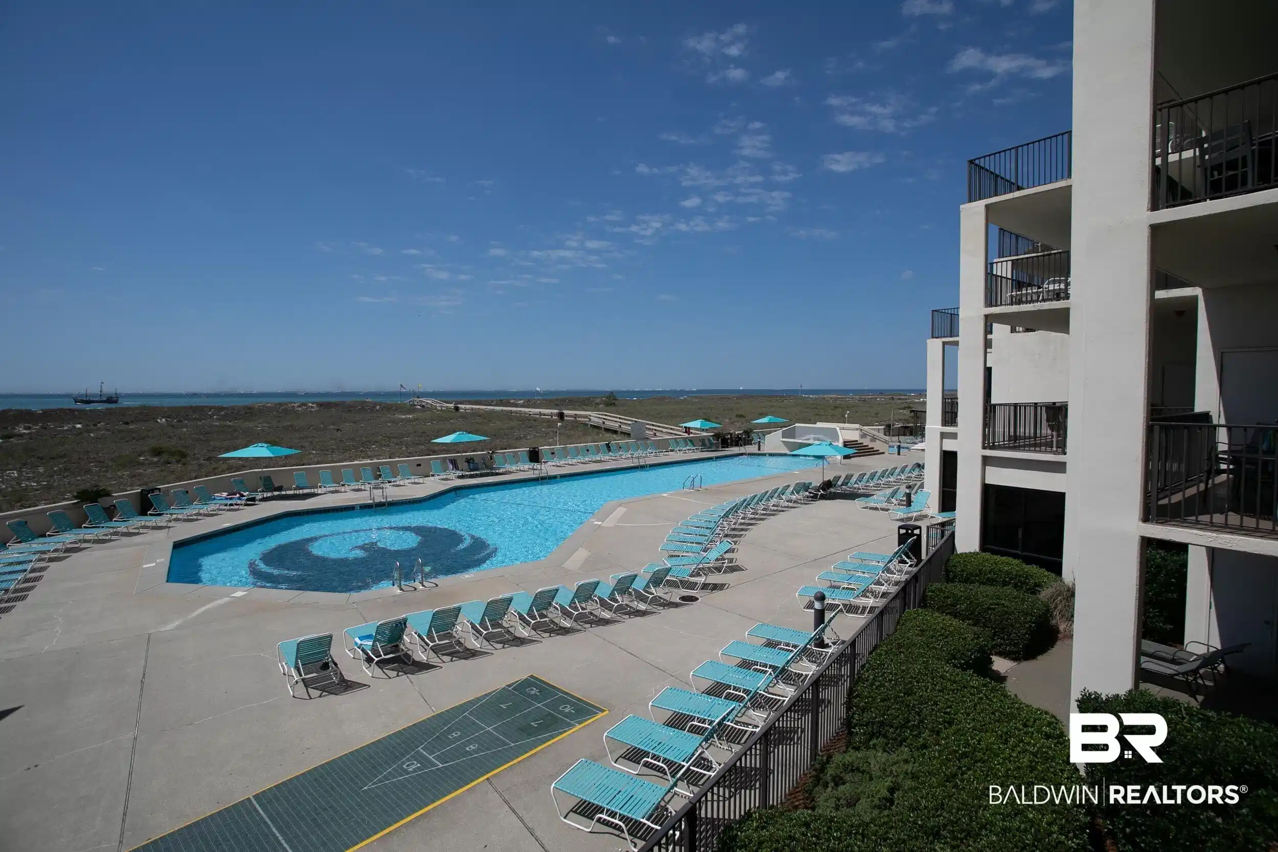 Image 1: Outdoor swimming pool with integrated design element, surrounded by an expansive concrete deck, numerous lounge chairs, and a shuffleboard court