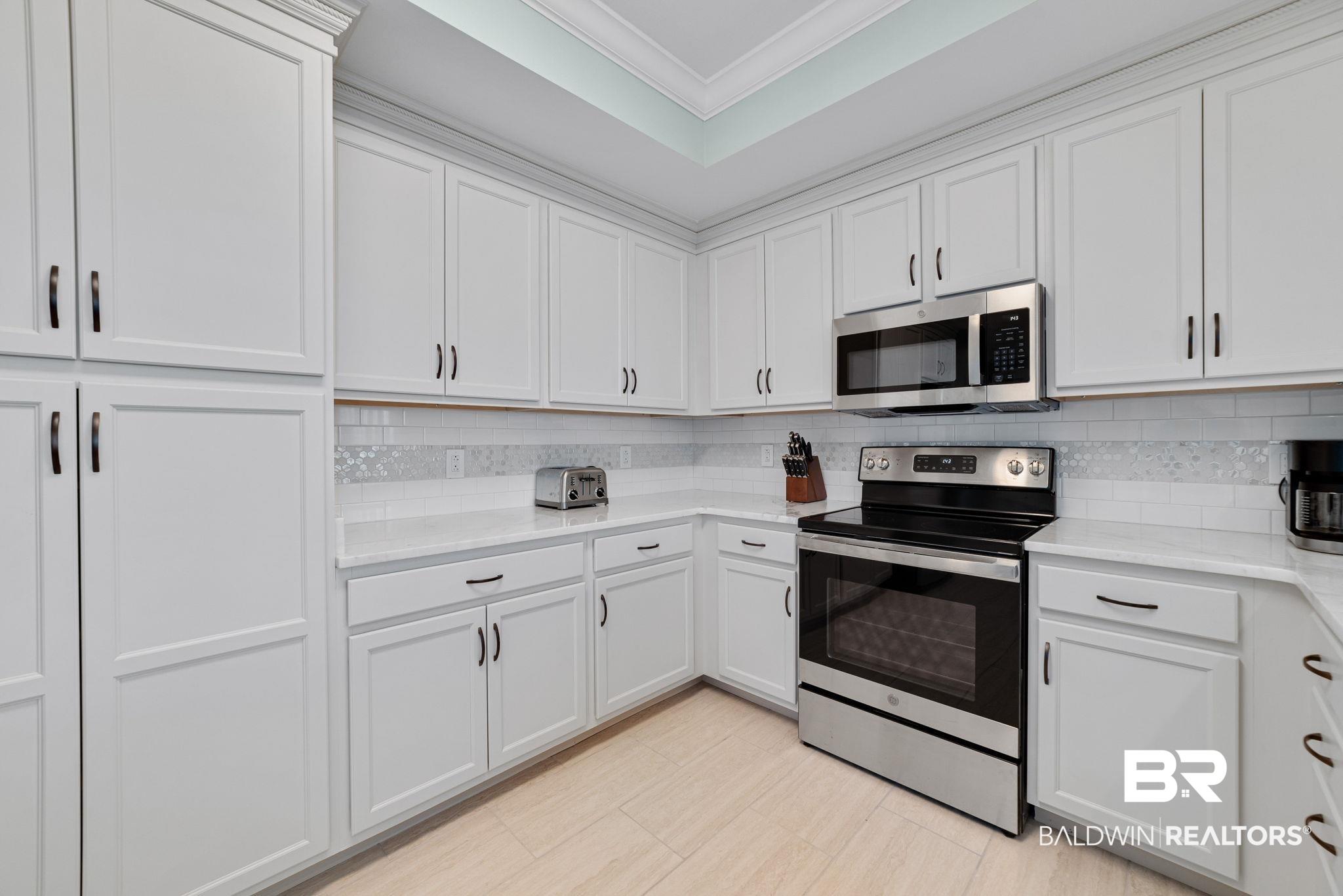 Image 4: Kitchen featuring extensive white cabinetry with black hardware, stainless steel appliances, a tile backsplash with decorative accents, and light-toned tile flooring