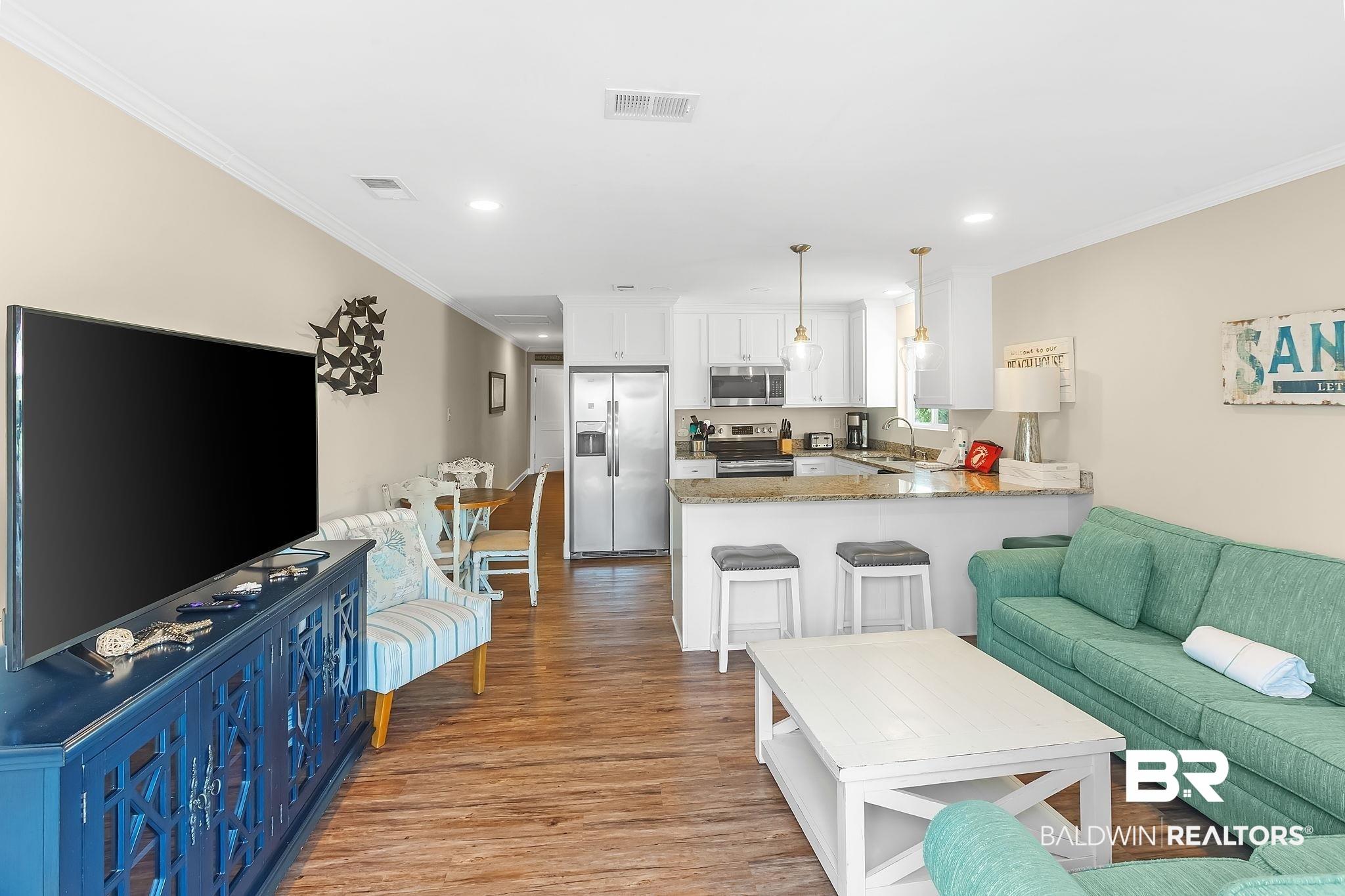 Image 2: Open-concept living space featuring wood-finish flooring, a kitchen with white cabinetry, stainless steel appliances, and a breakfast bar with pendant lighting
