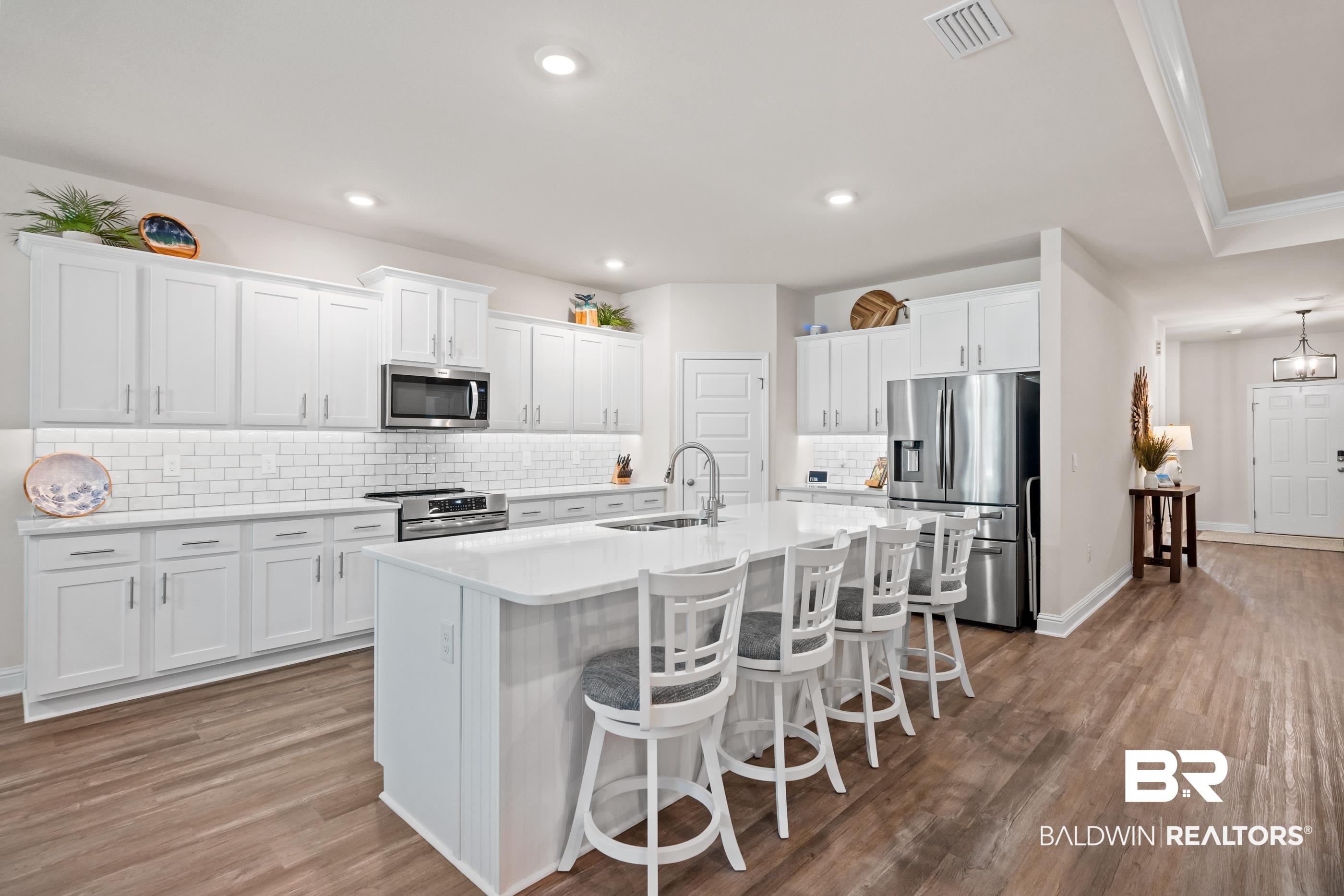 Image 3: Spacious kitchen featuring white shaker-style cabinetry, a large center island with seating, stainless steel appliances, a subway tile backsplash, and wood-finish flooring