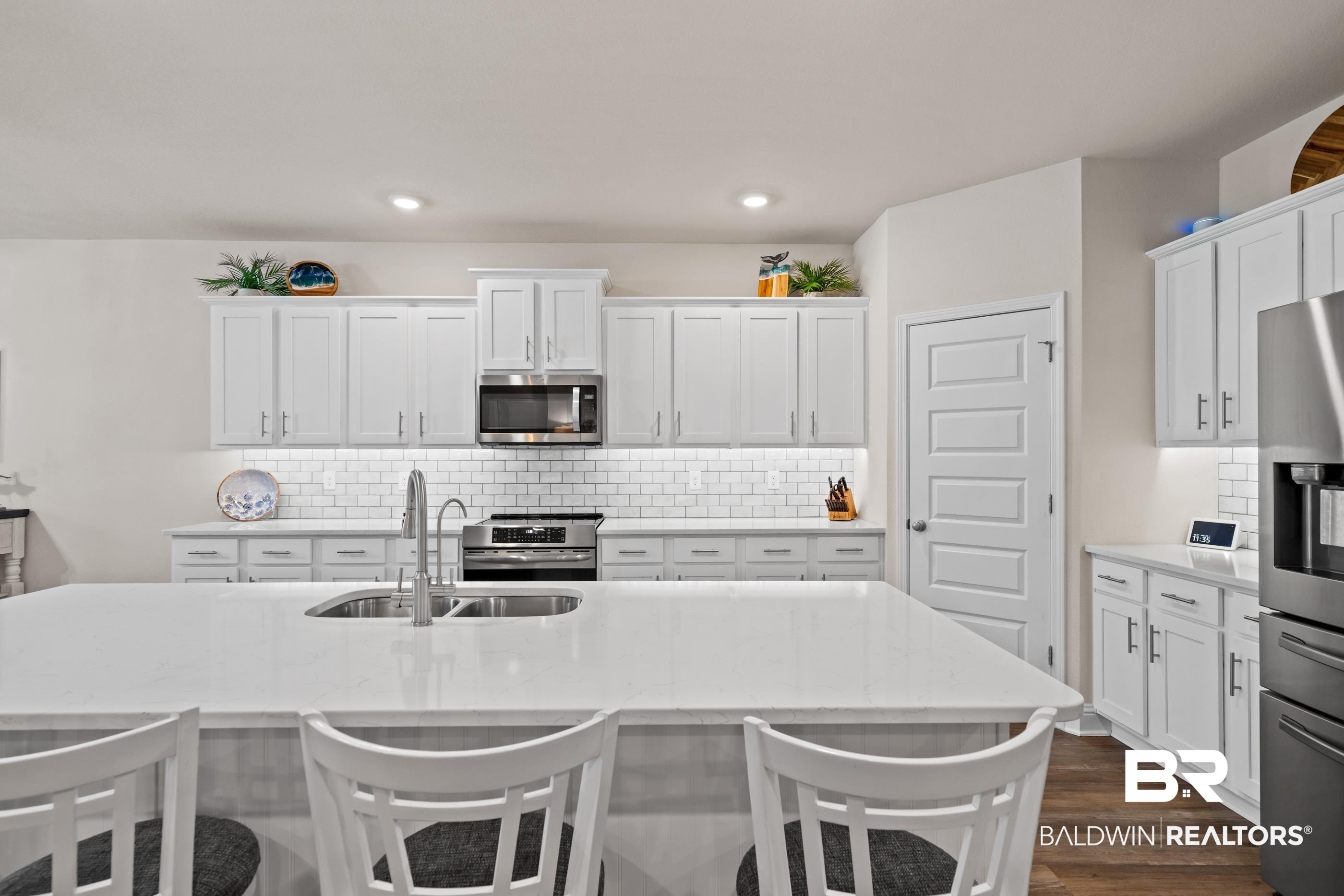 Image 4: Spacious kitchen featuring an expansive central island with an undermount double-basin sink, white shaker-style cabinetry, subway tile backsplash, stainless steel appliances, and recessed lighting