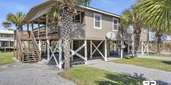 Image 2: Raised coastal-style home featuring a covered porch with wood railing, exterior stairs, and gravel driveway