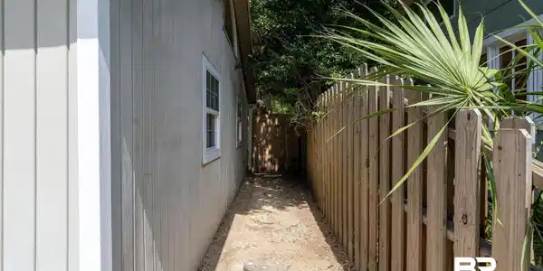Image 3: Board and batten siding with white-trimmed windows, a wooden picket fence, and established palm foliage