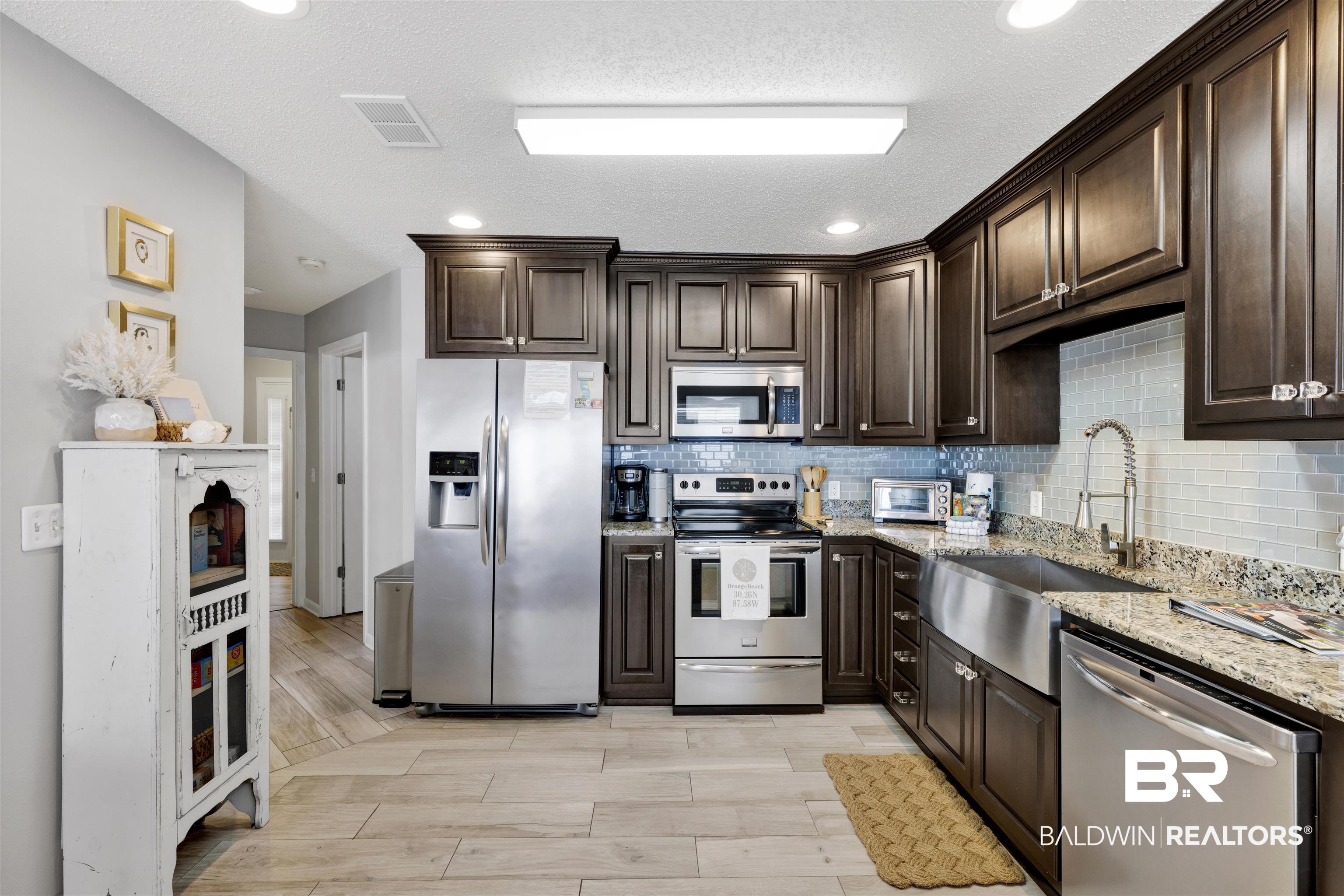Image 3: Kitchen featuring dark wood finish cabinetry, stainless steel appliances, light stone countertops, wood finish floors, and a textured ceiling
