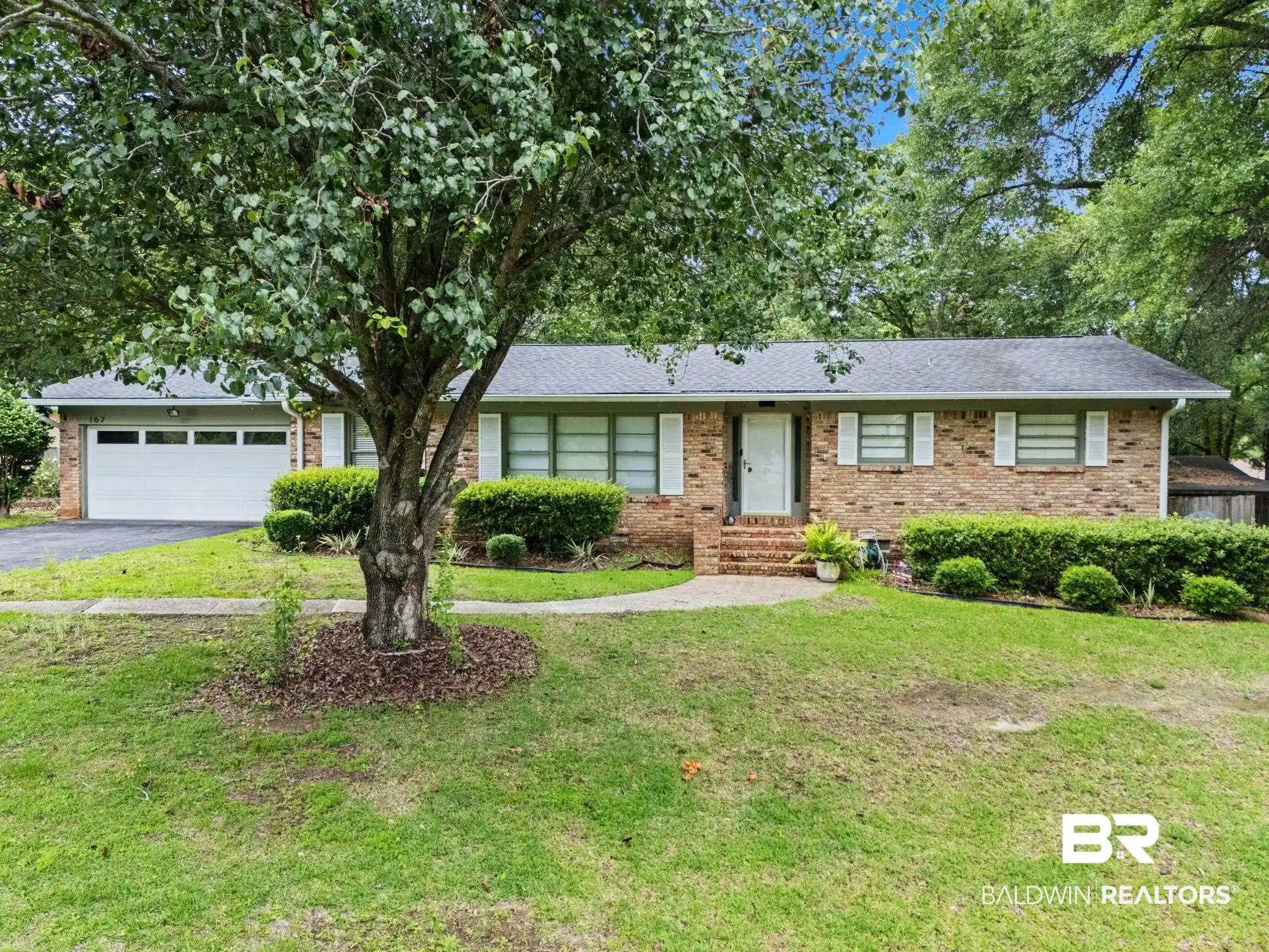 Image 1: Brick-sided residence featuring a prominent mature tree in the front yard, a white paneled garage door, a front entry with brick steps, a dark shingle roof, and white-framed windows with decorative shutters
