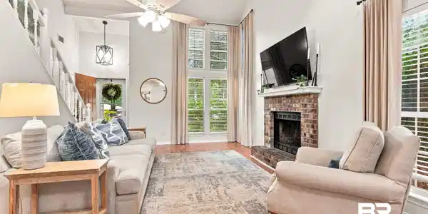 Image 4: Spacious living area featuring a vaulted ceiling with ceiling fan, wood-finish flooring, a brick fireplace with a white mantel, and tall windows with blinds
