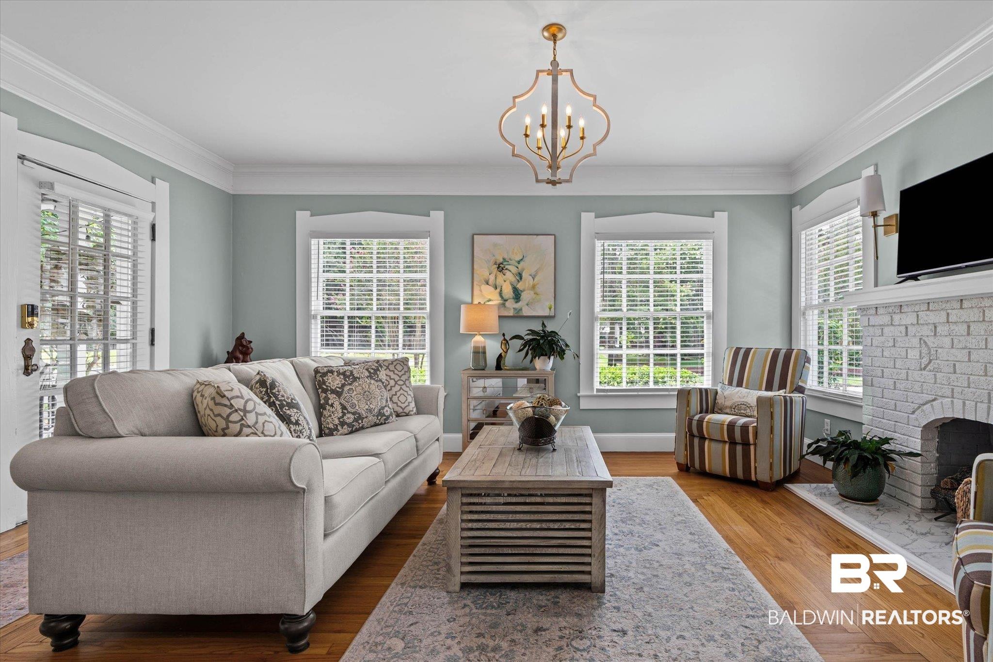 Image 4: Spacious living area featuring warm wood-finish flooring, a white-painted brick fireplace with a mantel, and multiple windows with white blinds