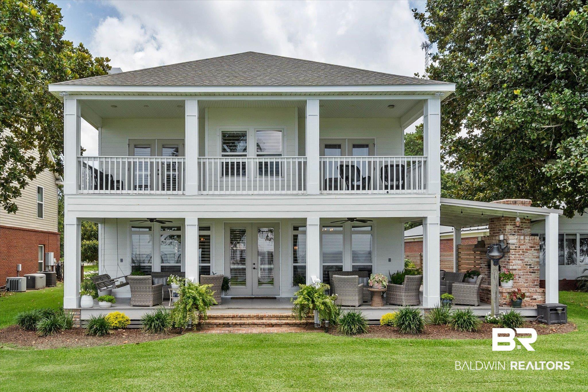 Image 1: Two-story waterfront exterior with expansive covered porch, second-story balcony, brick accent wall, double French doors, and manicured lawn