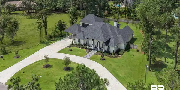 Image 2: Expansive property featuring a white exterior with dark shutters, a multi-gabled dark roof, a covered entry porch with columns, a curved concrete driveway, and mature trees surrounding the perimeter