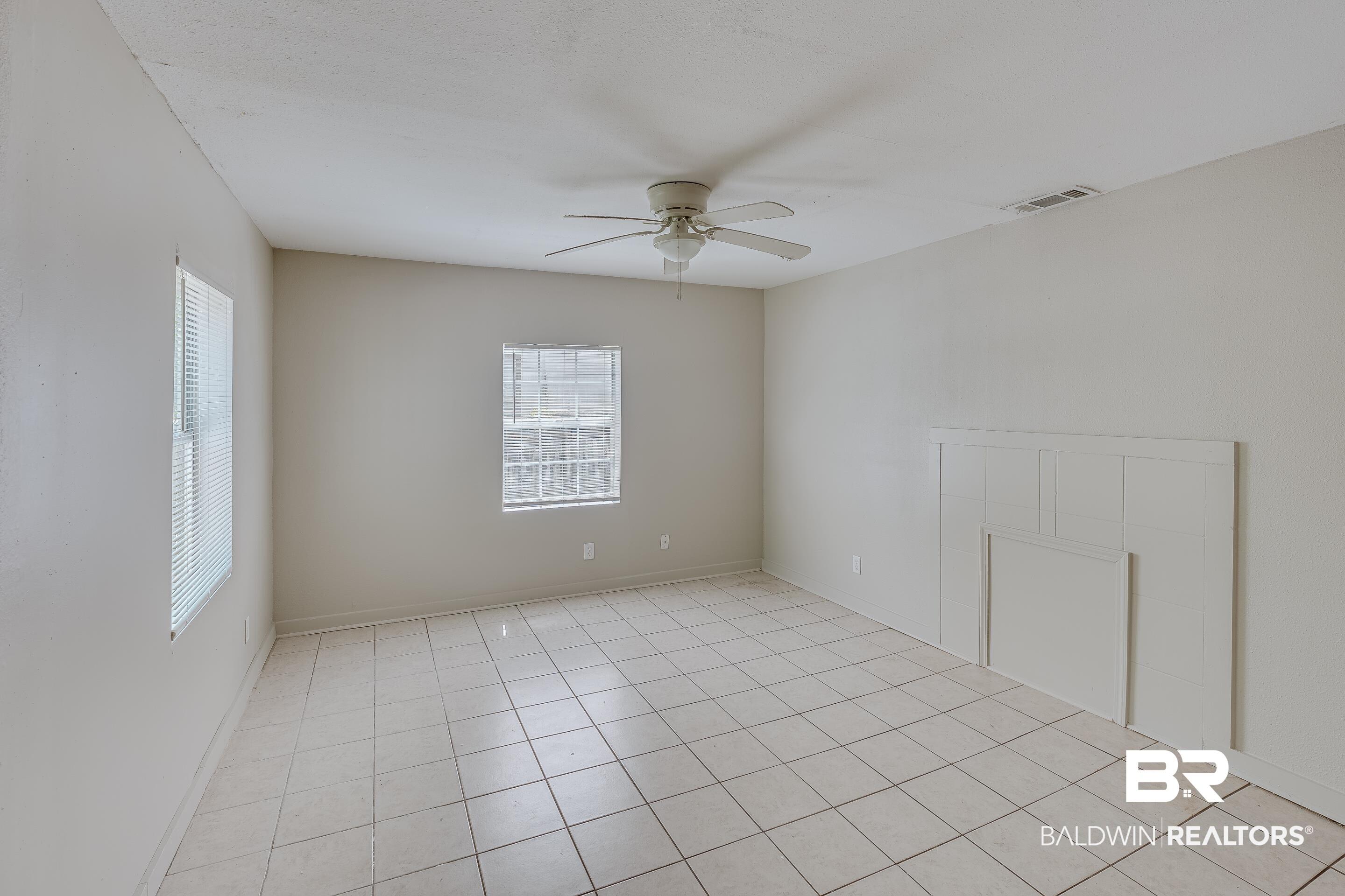 Image 3: Room featuring light-toned tile flooring, two windows with blinds, a ceiling fan, and a decorative wall panel accent
