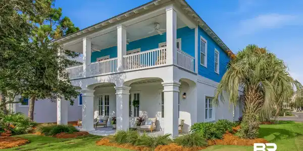 Image 3: Two-story coastal-style residence featuring a prominent covered porch, upper-level balcony, white columns, blue siding accents, and mature landscaping