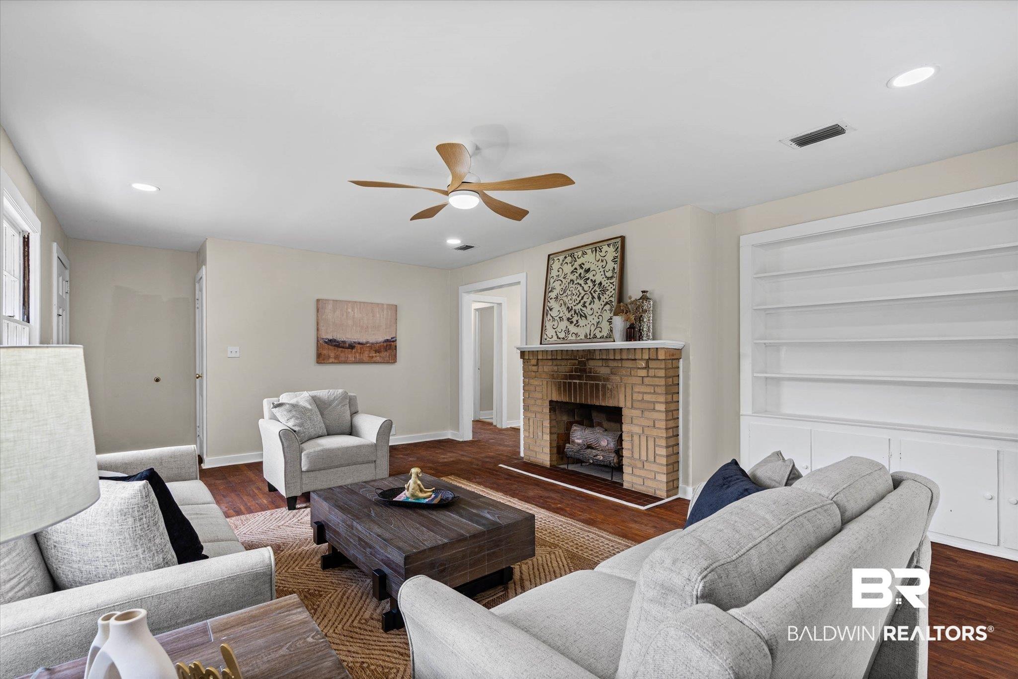 Image 3: Living area featuring warm wood-finish flooring, a brick fireplace with mantel, and built-in shelving
