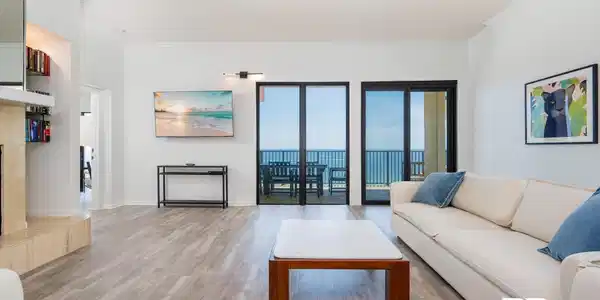 Image 2: Spacious living area featuring light wood-finish flooring, a built-in shelving unit, a wall-mounted flat-screen television, and expansive sliding glass doors opening to a balcony with water scenery