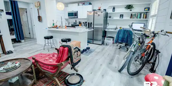 Image 4: Open-concept living area featuring light wood-finish flooring, white shiplap-style wall accents, a kitchen with white cabinetry and stainless steel appliances, a breakfast bar, and integrated shelving