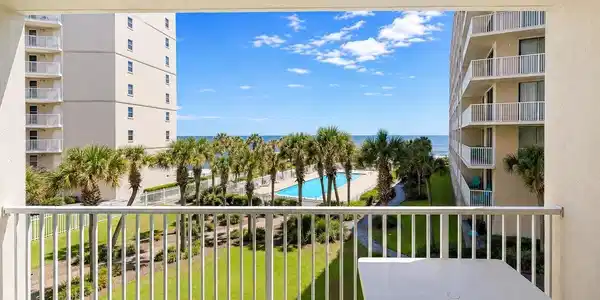 Image 3: Balcony with white metal railing overlooking a landscaped lawn with palm trees, a swimming pool, and the ocean beyond