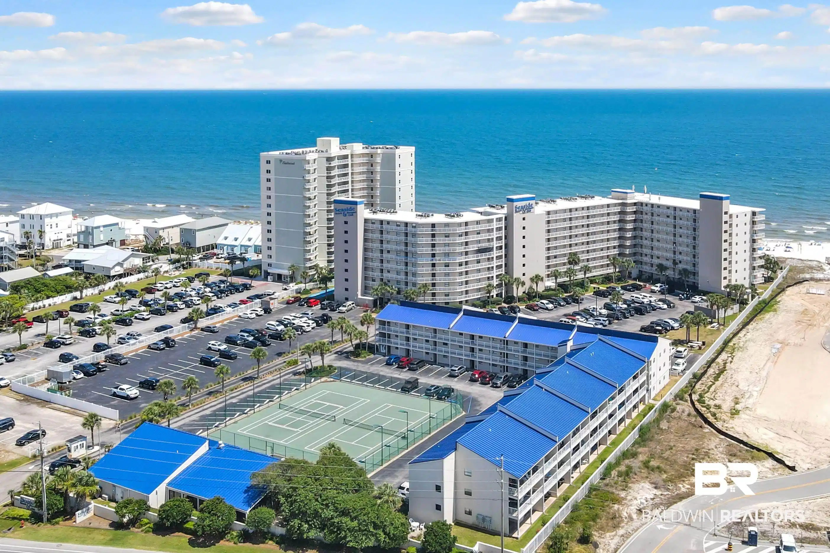 Image 1: Coastal property with multiple buildings featuring a tennis court, expansive parking areas, and convenient beach access