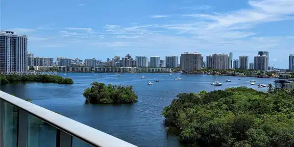 Image 4: The balcony captures wide water views with surrounding greenery, boats, and the city skyline beyond.