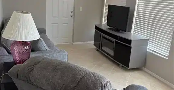 Image 4: Living area featuring a white entry door with an arched glass insert, two windows with horizontal blinds, light tile flooring, and light gray wall paint