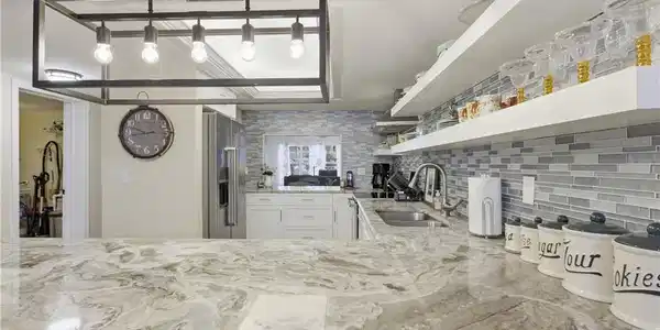 Image 4: Spacious kitchen featuring a large island with a natural stone countertop, a modern rectangular pendant light fixture, a mosaic tile backsplash, white cabinetry, and stainless steel appliances