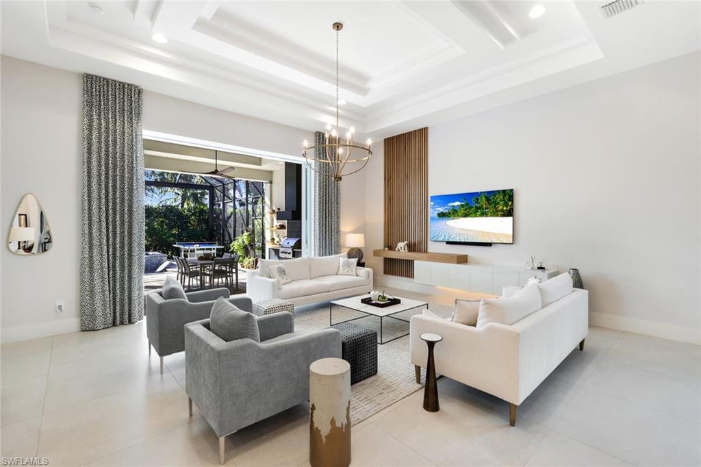 Image 4: Spacious living area featuring high ceilings with tray detailing and recessed lighting, large format tile flooring, a custom accent wall with integrated wood-finish paneling and floating cabinetry, and expansive sliding glass doors opening to an outdoor living space with a screened enclosure