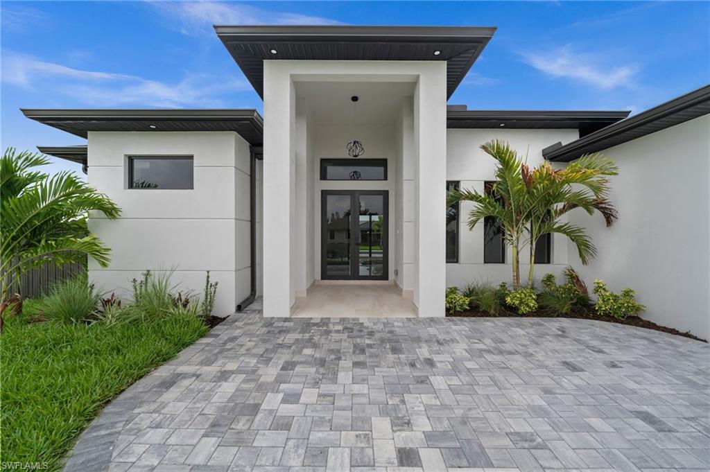 Image 4: Contemporary exterior featuring a recessed entry, dark-framed double glass doors, a paver driveway, white stucco finish, and dark fascia with integrated lighting
