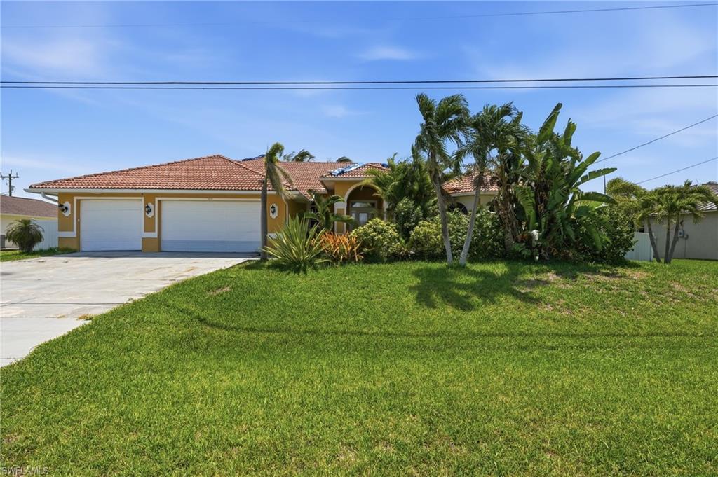 Image 1: Yellow stucco exterior featuring a terra cotta tile roof, two white garage doors, a concrete driveway, and established tropical landscaping